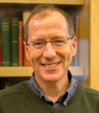 a smiling white man in glasses and a green sweater in front of bookshelves